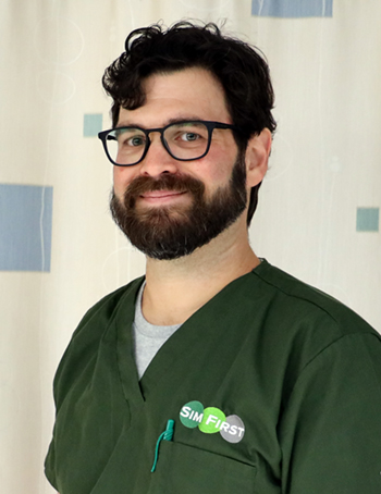 A bearded man in black glasses and olive green medical scrubs