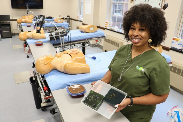 A woman wearing navy green medical scrubs standing in a simulation laboratory, holding a simulation trainer unit