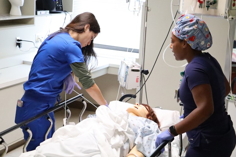 A woman in royal blue medical scrubs and a woman in navy blue medical scrubs examining a simulation manikin