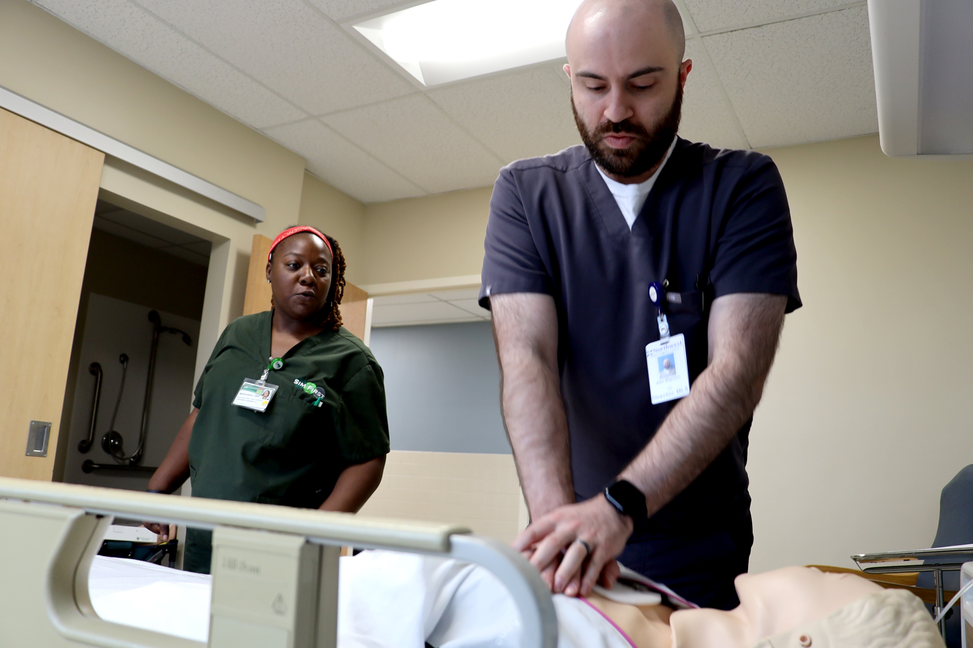 A woman in green medical scrubs instructs a man in gray medical scrubs in a simulation exercise.