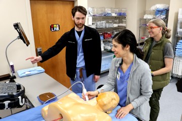 Two women and one man, all wearing medical scrubs and participating in a procedural clinical simulation with a manikin and ultrasound