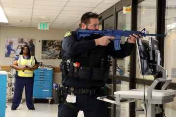 A male police officer holding a prop assault rifle in a hospital hallway, as a woman wearing a safety vest observes in the background