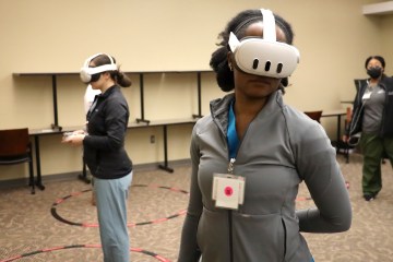 Two women in medical scrubs and jackets wearing virtual reality headsets and participating in a VR simulation with a female simulation technician in the background
