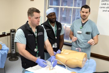 Three men in medical scrubs simulating ultrasound technique on a simulation manikin