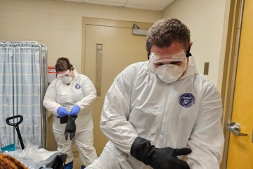 Two health-care workers, one male and one female, fastening black gloves over their white hazardous material suits