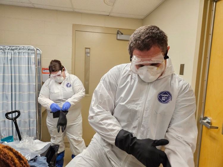 Two health-care workers, one male and one female, fastening black gloves over their white hazardous material suits