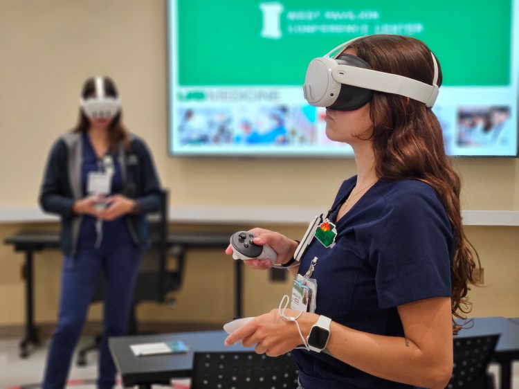 Two Caucasian women in navy blue scrubs wearing white virtual reality masks and holding white virtual reality controllers