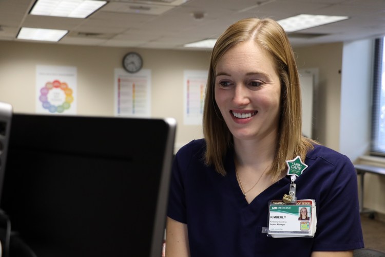 A Caucasian woman with blond hair wearing navy blue medical scrubs smiling in front of a computer monitor during a health-care simulation