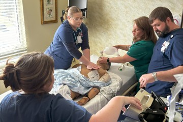 Two female nurses in navy blue scrubs, a female nurse in green scrubs and a male nurse in navy blue scrubs interacting with a medical simulation manikin