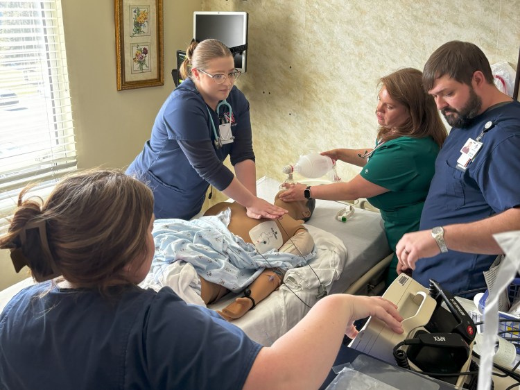 Two female nurses in navy blue scrubs, a female nurse in green scrubs and a male nurse in navy blue scrubs interacting with a medical simulation manikin