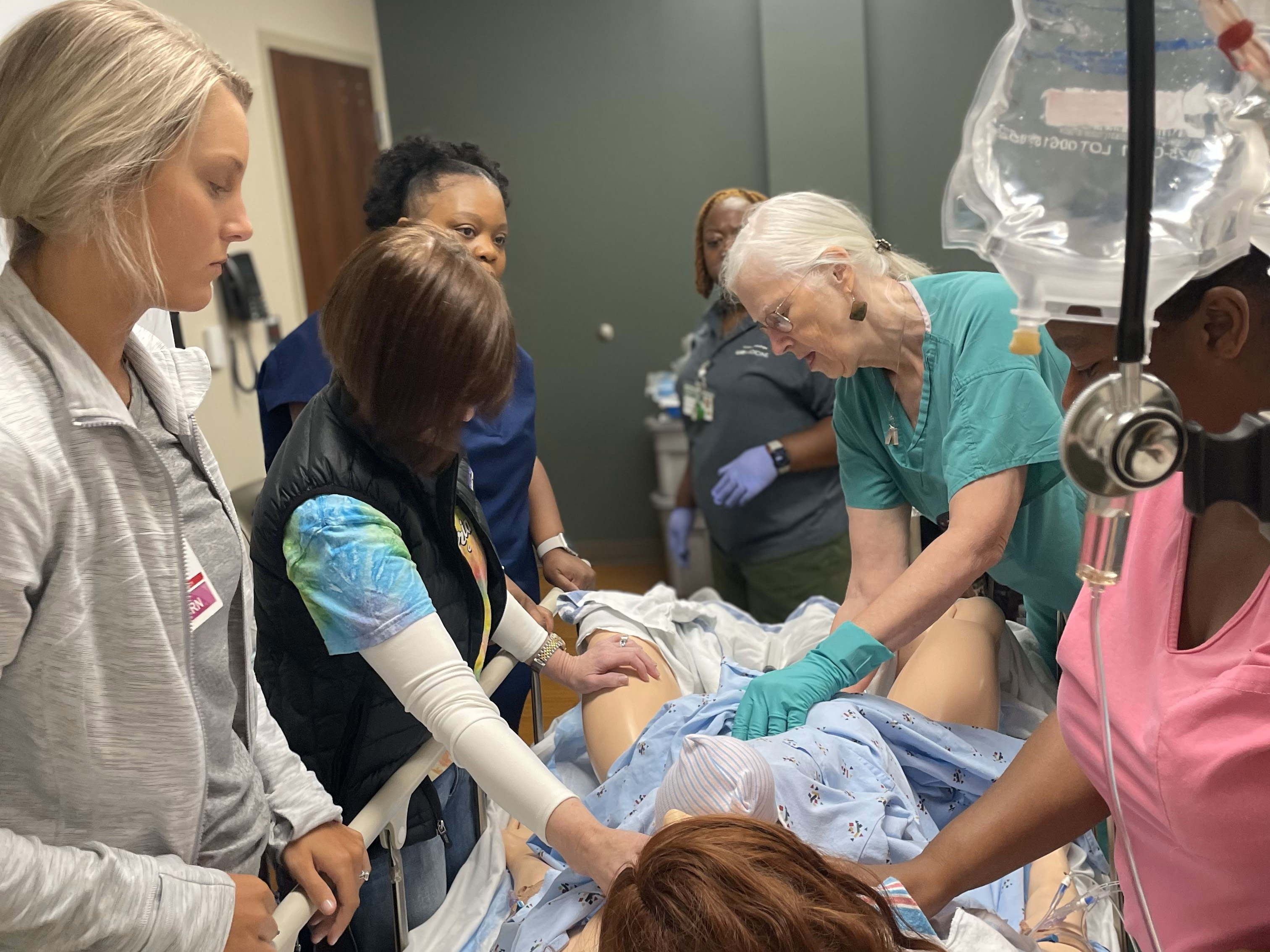 Five female health-care workers in scrubs of varying colors observing a female instructor in green scrubs demonstrating OB practices on a medical manikin