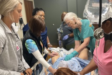 Five female health-care workers in scrubs of varying colors observing a female instructor in green scrubs demonstrating OB practices on a medical manikin