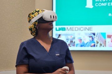 A female nurse in navy blue scrubs wearing a white virtual reality headset and holding a white virtuality headset and a white virtual reality hand control.
