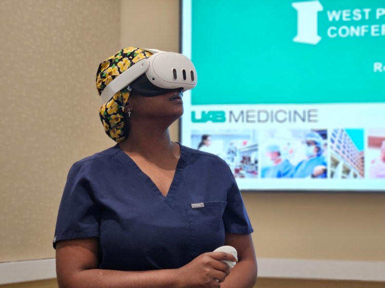 A female nurse in navy blue scrubs and a scrub cap with a yellow floral pattern wearing a white virtual reality headset and holding a white virtuality headset and a white virtual reality hand control.