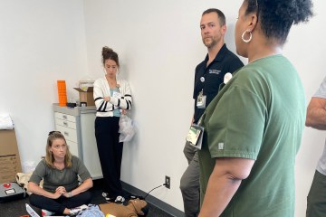 A Black woman in olive green medical scrubs leading a health-care simulation with a Caucasian man and two Caucasian women.