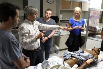 Three Caucasian men and a woman standing near a health-care simulation manikin in a simulated hospital room.