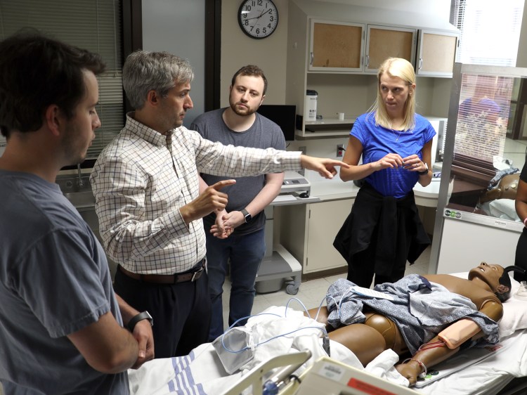 Three Caucasian men and a woman standing near a health-care simulation manikin in a simulated hospital room.