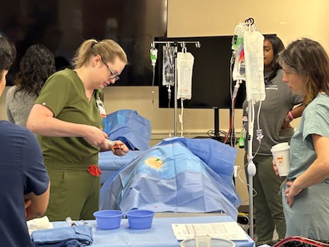 A female health-care professional in olive green scrubs practicing cardiac catheterization on a simulation manikin with a female facilitator in light blue scrubs overseeing