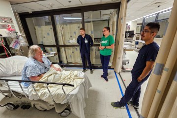 An elderly Caucasian woman wearing a medical gown in a hospital bed animatedly addressing a Caucasian male nurse wearing navy blue scrubs, a Caucasian female nurse wearing a green T-shirt and navy blue scrub bottoms and a Latin American male nurse wearing navy blue scrubs