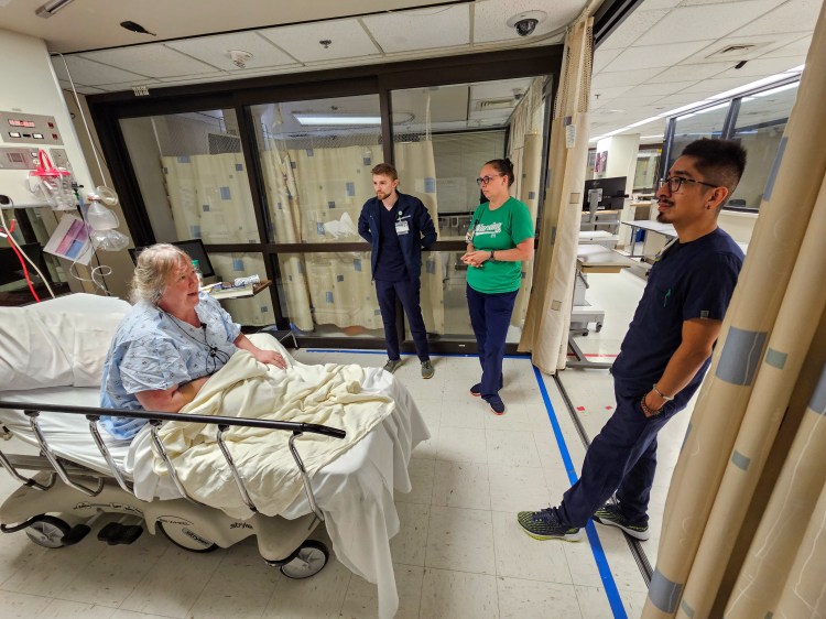 An elderly Caucasian woman wearing a medical gown in a hospital bed animatedly addressing a Caucasian male nurse wearing navy blue scrubs, a Caucasian female nurse wearing a green T-shirt and navy blue scrub bottoms and a Latin American male nurse wearing navy blue scrubs