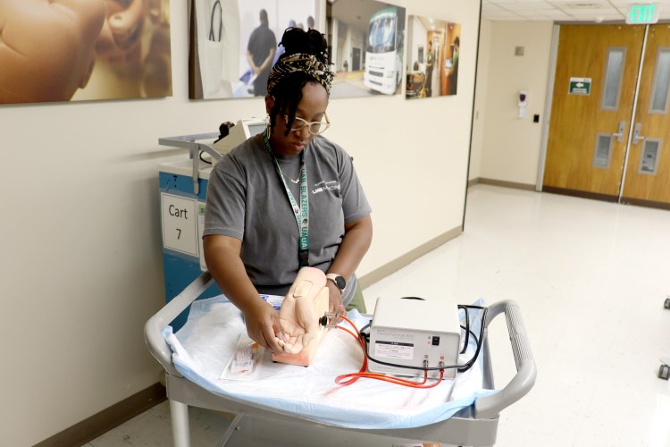 A Black female in a dark gray UAB Clinical Simulation T-shirt and olive green scrub bottoms preparing a simulation trainer shaped like a human hand for an equipment loan