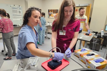 A Caucasian woman in light blue medical scrubs leading a Caucasian woman in burgundy medical scrubs through a simulated medical procedure