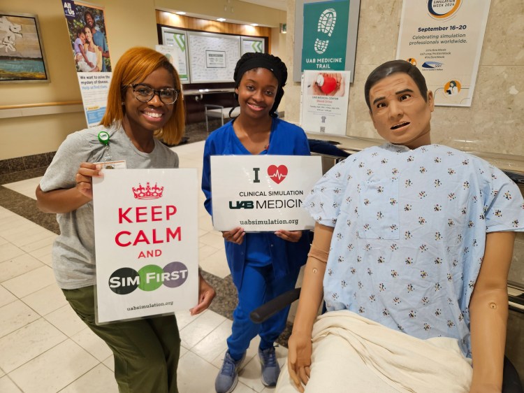 A Black woman in a heather gray T-shirt and olive green scrub pants and a Black woman in royal blue medical scrubs, both holding photo booth signs while posing next to a health-care simulation manikin