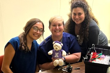 Three Caucasian women, two in navy blue medical scrubs and the third in gray medical scrubs, posing with a tabletop health-care simulation