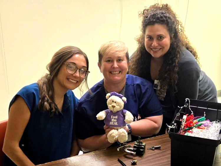 Three Caucasian women, two navy blue medical scrubs and the third in gray medical scrubs, posing with a tabletop health-care simulation