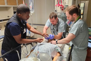 One Black man in navy blue medical scrubs and two Caucasian men in light blue medical scrubs simulating critical care support on a health-care simulation manikin