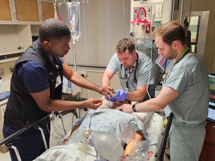 One Black man in navy blue medical scrubs and two Caucasian men in light blue medical scrubs simulating critical care support on a health-care simulation manikin