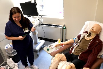 A Caucasian woman in navy blue medical scrubs searching for simulated patient safety errors in a simulated hospital room with a health-care simulation manikin