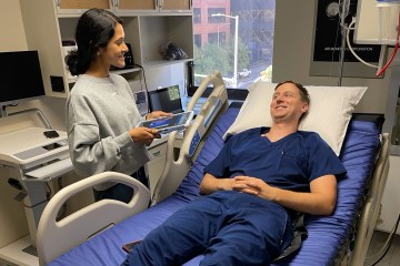 A female physician in medical scrubs and a gray sweatshirt talking with a male physician in navy blue medical scrubs laying on a hospital bed as part of a health-care simulation