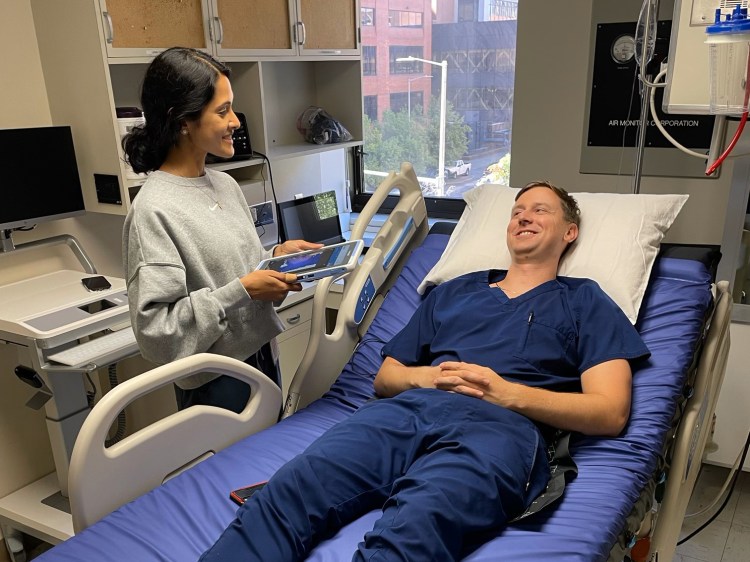 A female physician in medical scrubs and a gray sweatshirt talking with a male physician in navy blue medical scrubs laying on a hospital bed as part of a health-care simulation