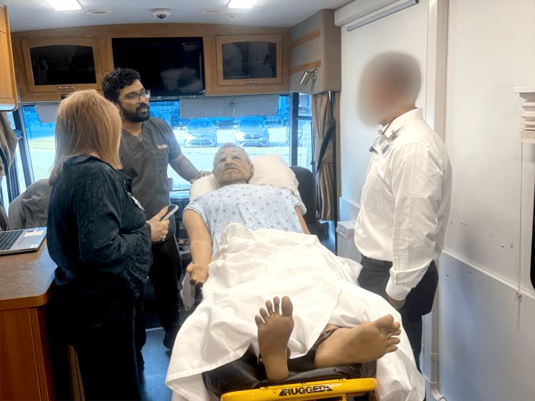 A man in olive green medical scrubs showcasing a health-care simulation manikin to other medical professionals inside of a mobile simulation lab