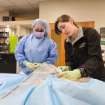 A woman in blue medical scrubs receiving instruction from a woman in a black jacket during a procedural central venous line insertion health-care simulation