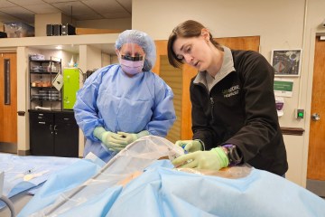 A woman in blue medical scrubs receiving instruction from a woman in a black jacket during a procedural central venous line insertion health-care simulation