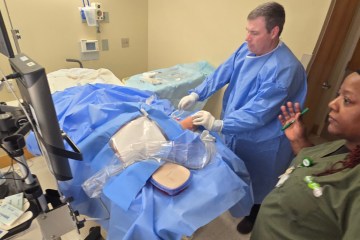 A man in a blue surgical gown performing a simulated medical procedure while standing next to a woman in olive green medical scrubs