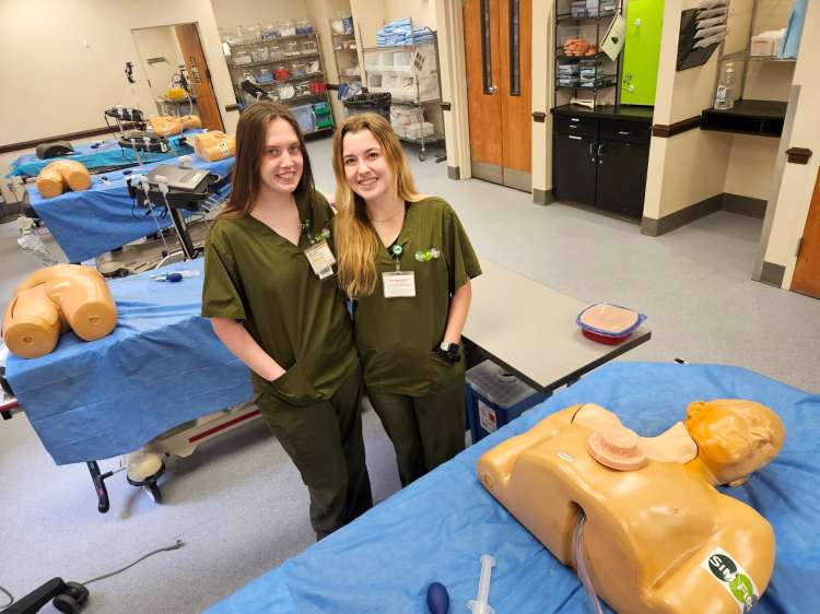 Two young Caucasian females in olive green medical scrubs standing next to health-care simulation manikins in a procedural simulation lab
