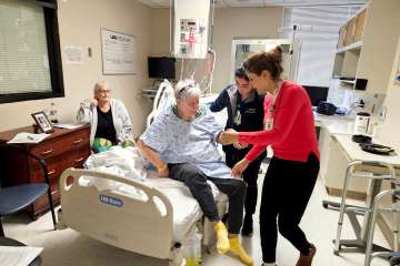 An elderly woman sitting next to an elderly man wearing a hospital gown, being assisted out of bed by two female medical professionals in medical scrubs and jackets