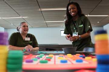 Two women in olive green medical scrubs setting up a tabletop health-care simulation