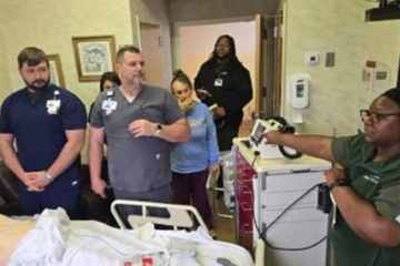 A woman and man in olive green medical scrubs facilitating a health-care simulation for four health-care professionals in multi-colored medical scrubs