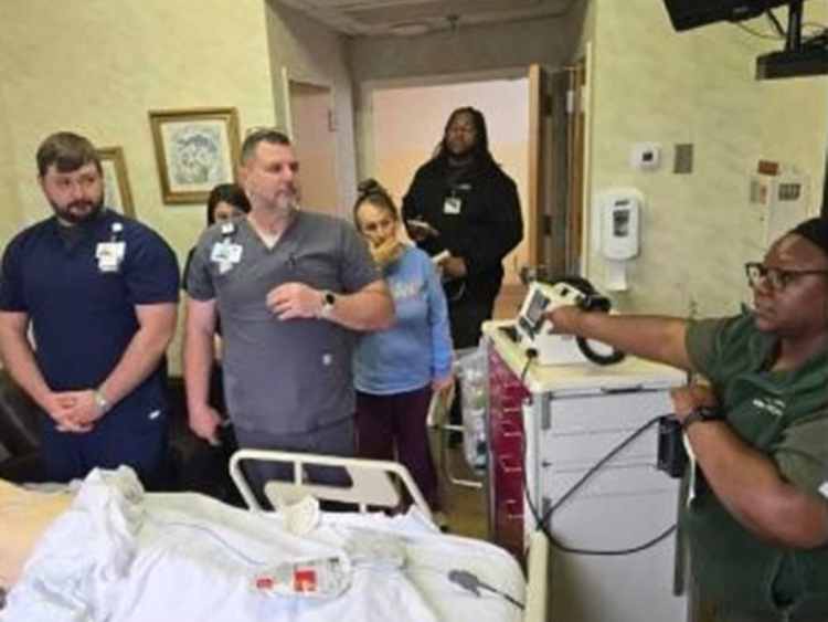 A woman and man in olive green medical scrubs facilitating a health-care simulation for four health-care professionals in multi-colored medical scrubs