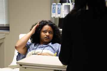 A woman portraying a patient in a hospital bed conversing with a person in the foreground