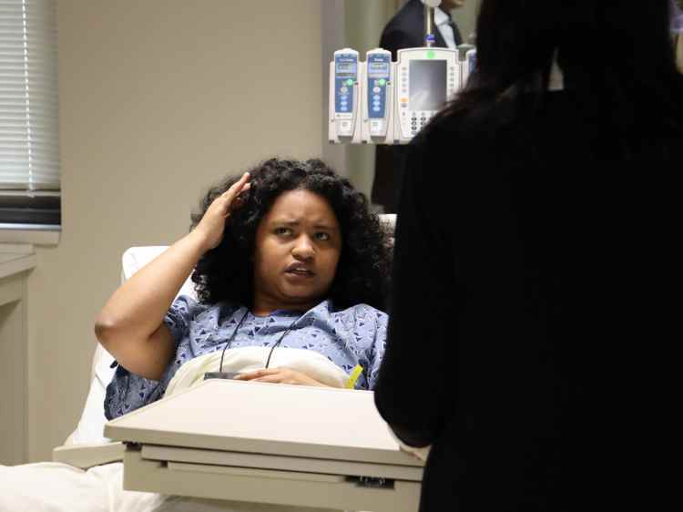 A woman portraying a patient in a hospital bed conversing with a person in the foreground