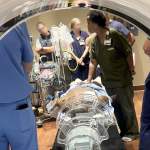 A woman in olive green medical scrubs speaking with health-care professionals as seen through an opening in a medical device