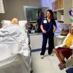 Two women in navy blue medical scrubs interacting with a female standardized patient and a high-fidelity health-care simulation manikin as part of a health-care simulation
