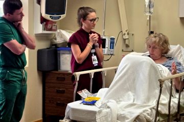 Two health-care professionals—a man in green medical scrubs and a woman in burgundy medical scrubs—interacting with a female standardized patient portraying a disruptive patient in a health-care simulation.