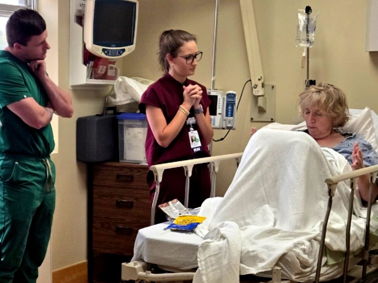 Two health-care professionals—a man in green medical scrubs and a woman in burgundy medical scrubs—interacting with a female standardized patient portraying a disruptive patient in a health-care simulation.