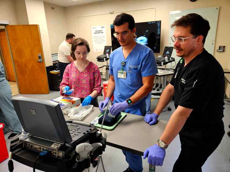 A woman in a floral blowse and two men, one in light blue medical scrubs and the other in black medical scrubs, participating in a health-care simulation with an ultrasound device